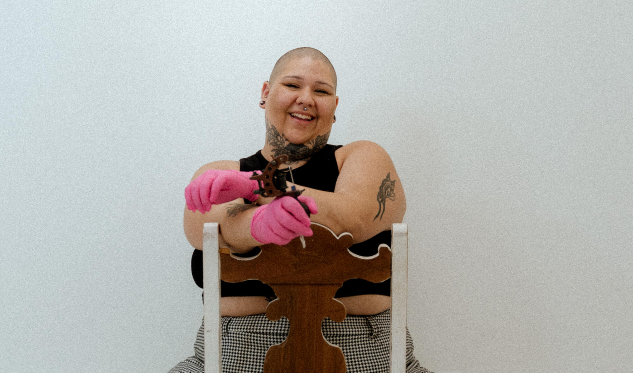 Toni PNW, a fat tattoo artist, sits in a chair smiling at the camera. She is wearing pink gloves and holding a tattoo machine.