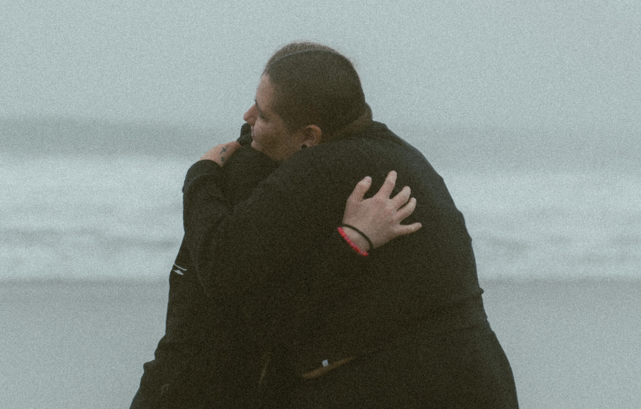 A thin woman and a fat woman hug one another at the beach on a cloudy day.